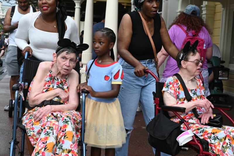 A young girl in a Snow White costume stands between two elderly women in wheelchairs, both wearing floral dresses and Minnie Mouse ears. They are surrounded by people walking in a busy outdoor setting.