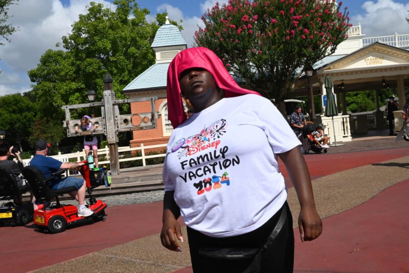 A person wearing a white shirt that reads "Disney Family Vacation 2024" stands outside. They have a pink cloth draped over their head. Other people and a building are visible in the background on a sunny day.