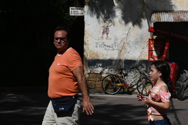 A man in an orange shirt and a girl with a floral shirt walk past a weathered wall with a faded cartoon drawing. A parked bicycle and a small refreshment booth are in the background, partially lit by sunlight.