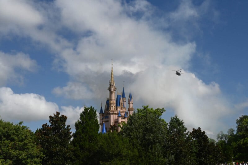 A tall, fairy tale castle with blue spires rises above green trees under a cloudy blue sky. A helicopter is visible in the distance.