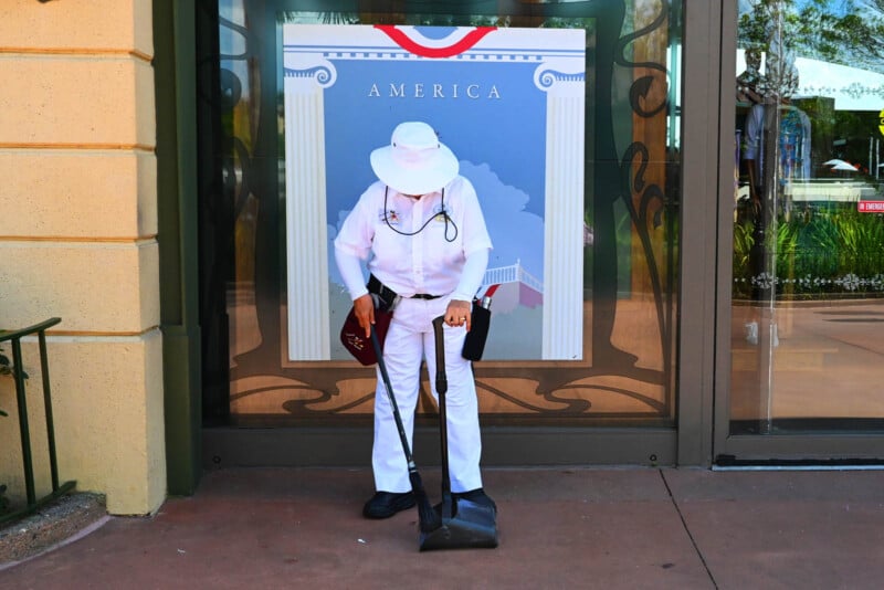 A person dressed in white, including a hat and shoes, bows their head while holding a black bag in front of an American-themed poster. The background features glass doors and decorative trim.