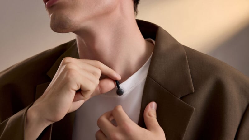 A person is adjusting a small black microphone clipped to a white shirt under a brown blazer. The focus is on the microphone and the person's hands.