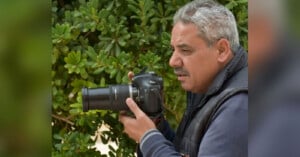 A man with gray hair and a mustache is holding a camera with a long lens, looking intently through it. He is wearing a dark vest and stands in front of lush green foliage.