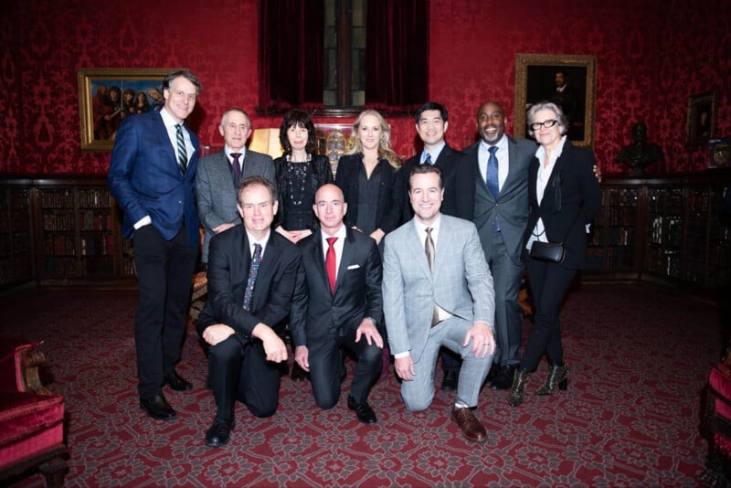 A group of ten people, dressed in formal attire, pose together in a richly decorated room with red walls and a patterned carpet. Bookshelves and framed paintings are visible in the background.