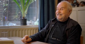 An elderly man with white hair and a beard is seated at a wooden table, smiling warmly. He's wearing a black jacket and has glasses hanging around his neck. In the background, there's a window with potted plants and a curtain.