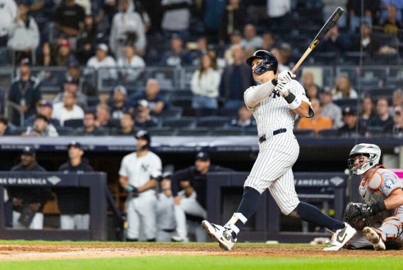 A baseball player in a pinstripe uniform swings a bat in a stadium. The batter watches the ball, while a catcher in gray and orange gear crouches behind. Spectators fill the stands in the background.