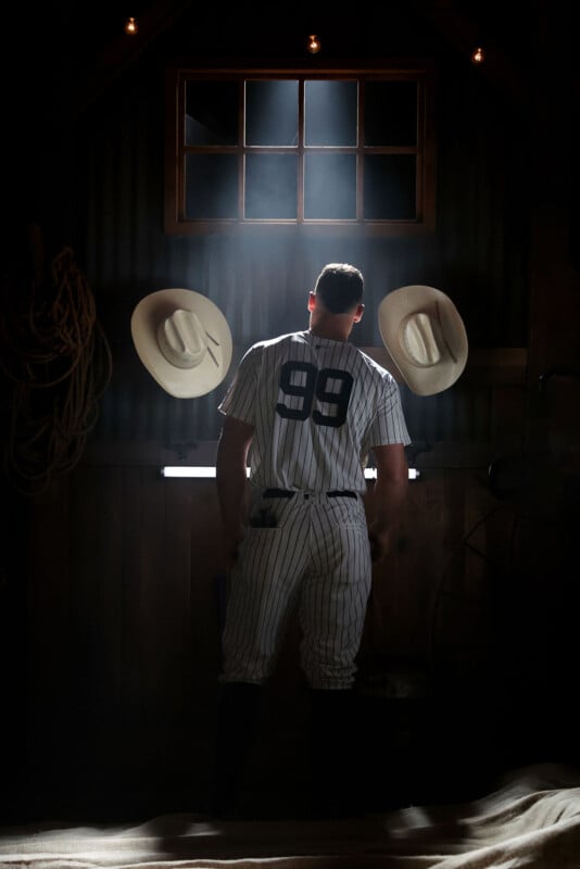 A baseball player wearing a pinstripe uniform with the number 99 stands in a dimly lit room, facing away. Two cowboy hats hang on the wall in front of him. Light filters in through a window above.