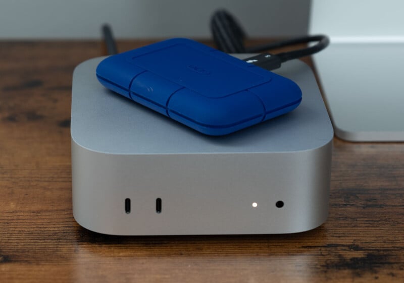 Silver desktop computer with a blue external hard drive on top, placed on a wooden desk. The computer has a few ports on its front panel and a small indicator light is illuminated. A black cable is connected to the hard drive.