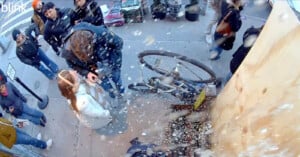 A man and woman stand on a sidewalk as a nearby construction barrier collapses. Dust and debris are scattering. A bicycle is knocked over. Nearby pedestrians react to the commotion while several items are visible on the street.