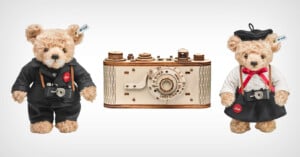 Two teddy bears dressed as photographers stand beside a vintage camera model. One bear wears a black suit and the other wears a black beret and white shirt. Each holds a miniature camera around their neck. The camera model is wooden with detailed features.