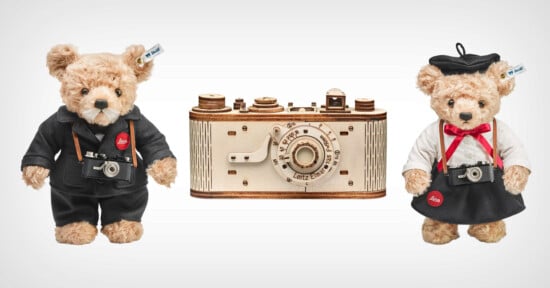 Two teddy bears dressed as photographers stand beside a vintage camera model. One bear wears a black suit and the other wears a black beret and white shirt. Each holds a miniature camera around their neck. The camera model is wooden with detailed features.