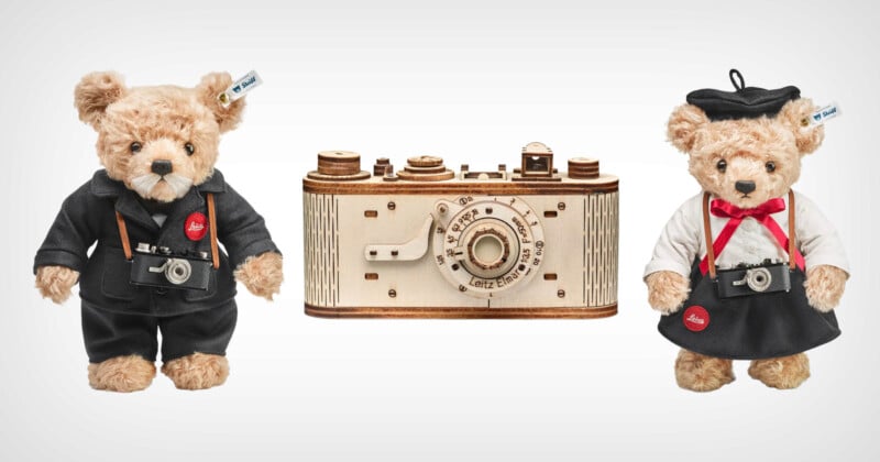 Two teddy bears dressed as photographers stand beside a vintage camera model. One bear wears a black suit and the other wears a black beret and white shirt. Each holds a miniature camera around their neck. The camera model is wooden with detailed features.