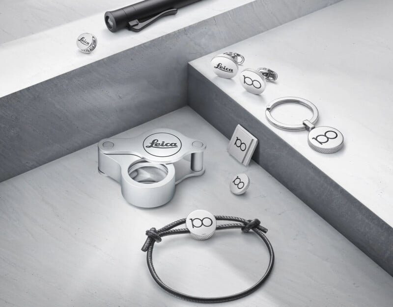 Modern Leica-branded accessories arranged on a minimalist concrete surface, including a flashlight, keychain, cufflinks, bracelet, and lens cap. Each item features the "Leica" logo, with some bearing a special "100" design.