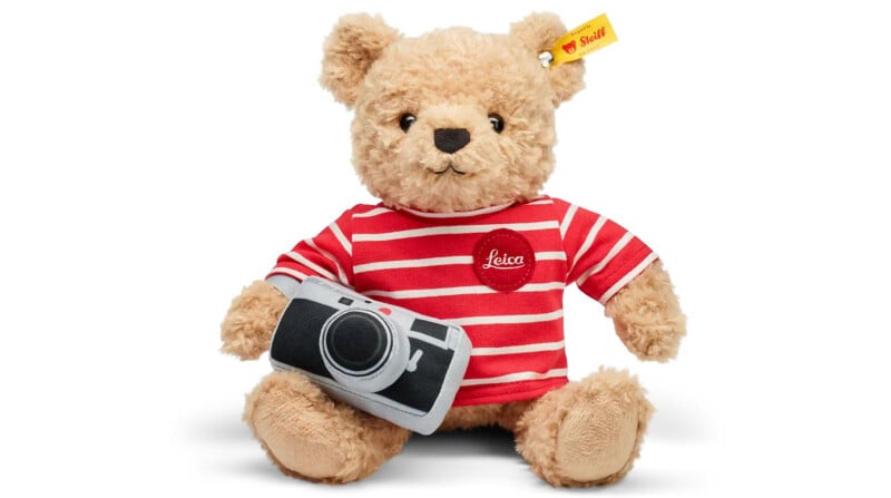 A plush teddy bear wearing a red and white striped shirt with a "Leica" logo. The bear is holding a soft toy camera. Its left ear has a small yellow tag with a red logo.