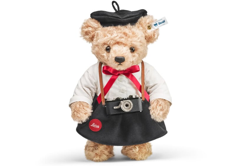 A teddy bear wearing a black beret and skirt, white shirt with a red bow tie, and a camera around its neck. The bear has a Steiff tag on its ear and a red Leica badge on the skirt.