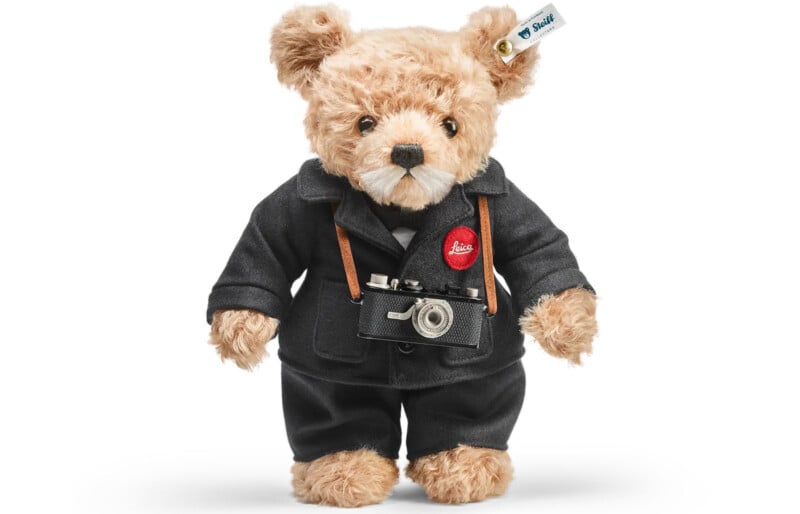 A plush teddy bear dressed in a black suit and bow tie, with a small black camera hanging around its neck. The bear's ear has a Steiff tag attached.