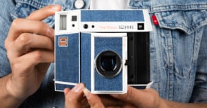 A person in a denim jacket holds a retro-style camera with a blue denim finish labeled "The Blues Square." The camera has a viewfinder, lens, and stylized branding resembling a vintage instant camera.