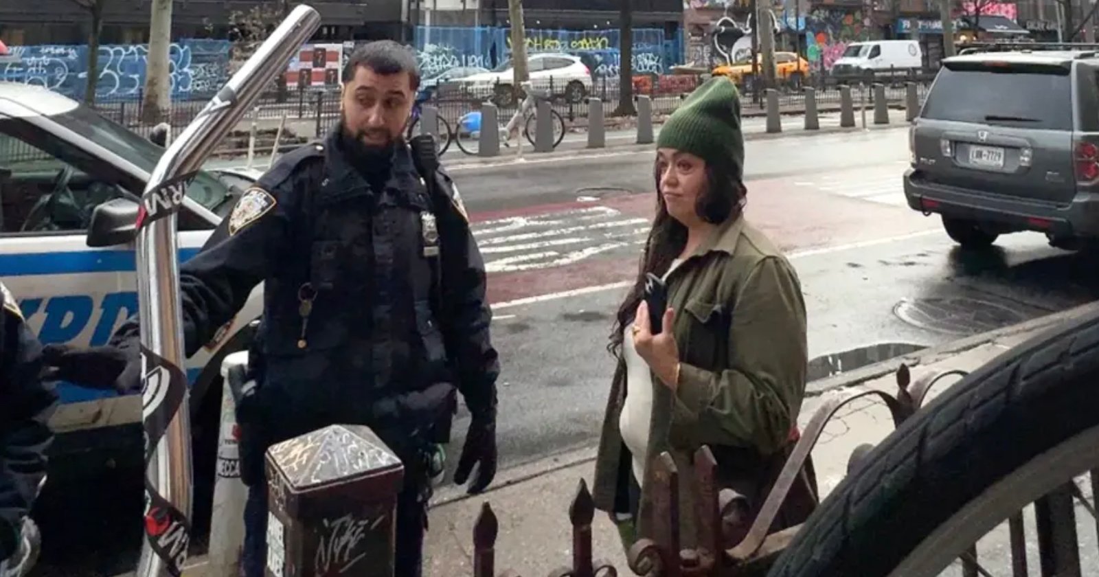 A police officer talks to a woman on a city sidewalk. The woman, wearing a green beanie, holds a bicycle frame. A police car is parked nearby, and graffiti is visible in the background.