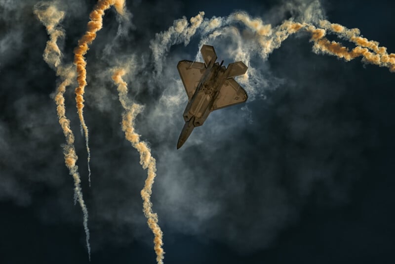 A military jet performs an acrobatic maneuver, leaving trails of smoke against a dark sky. The aircraft is angled downward with multiple swirling smoke trails adding dynamic movement to the scene.