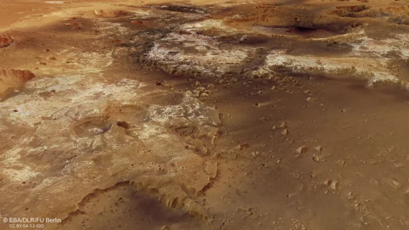 Aerial view of a Martian landscape with rocky terrain, featuring craters and elevated landforms. The surface appears reddish-brown with variations in texture and coloration.