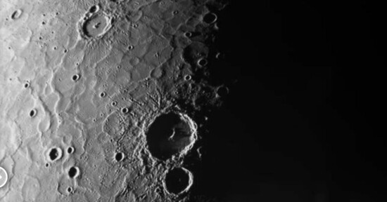 A detailed grayscale image of the Moon's surface, featuring numerous craters and a stark contrast between illuminated and shadowed areas. Several large craters are prominent in the center-left, highlighting the rugged lunar terrain.