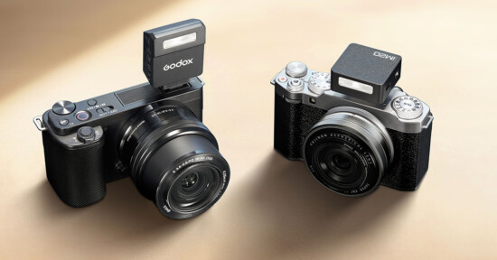 Two digital cameras are displayed, each equipped with a mounted external flash atop. The camera on the left has a "Godox" flash, while the camera on the right has an "AMED" flash. Both cameras have a sleek black and silver design.