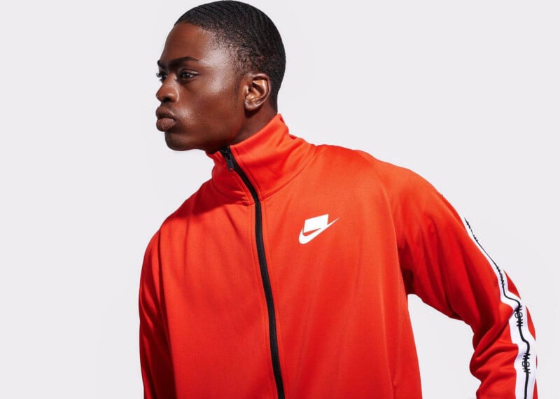 A person dressed in an orange Nike sports jacket featuring a white logo on the chest and black and white accents on the sleeves stands against a simple gray backdrop, gazing to the side.