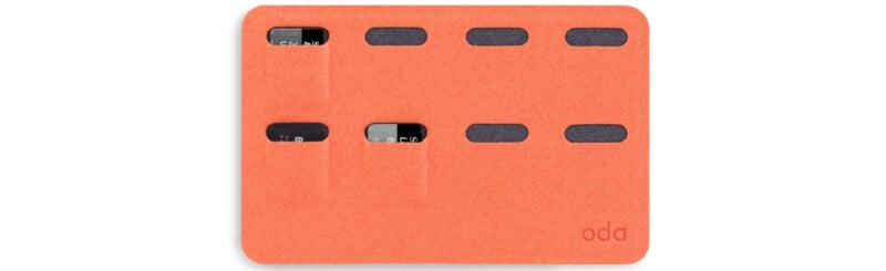 A slim, orange cardholder with the brand name "oda" in the bottom right corner. It features multiple horizontal slots with three partially visible black cards inserted. The cardholder has a minimalist design.