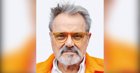 A man with gray hair and a beard wearing orange-rimmed glasses and a bright orange jacket looks directly at the camera against a plain background.