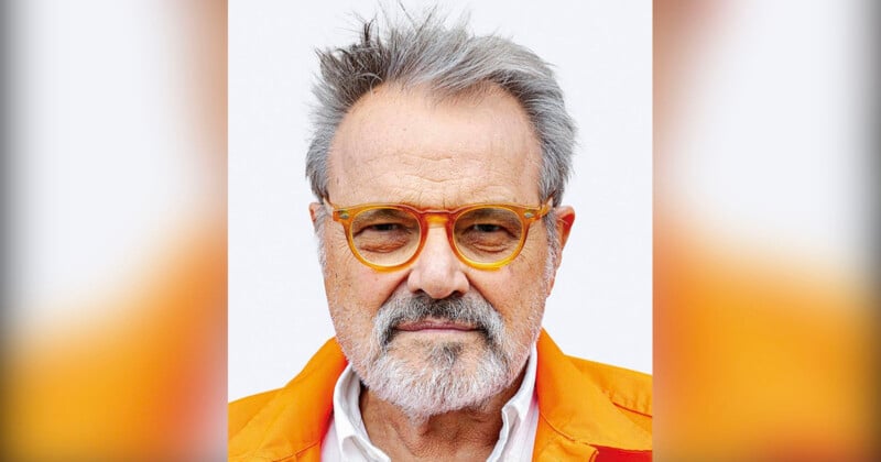 An elderly man with gray hair and a beard wears orange-rimmed glasses and a vivid orange jacket, gazing directly at the camera against a simple background.