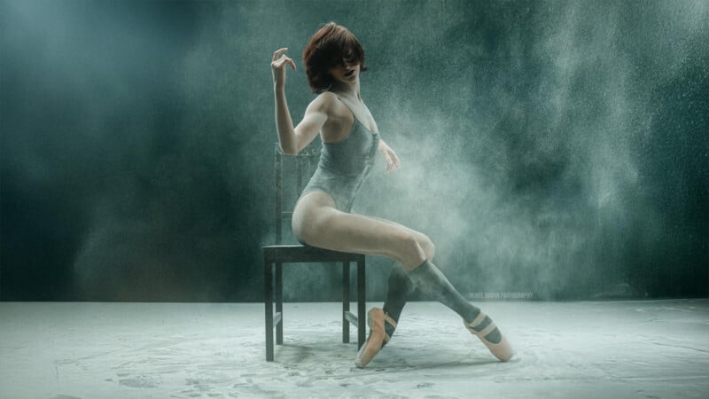 A dancer in a gray leotard performs an expressive pose while sitting on a chair. The background is filled with a cloud of dust or powder, creating a dynamic and artistic atmosphere. The setting is minimalist and moody.