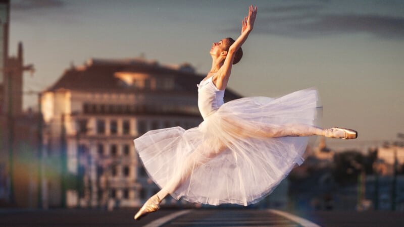 A ballerina in a white tutu leaps gracefully in mid-air on a city street at sunset. She is on pointe, her arms elegantly raised, with a blurred building in the background.