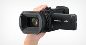 A person holding a black Panasonic 4K camcorder with a hexagonal lens hood and an LCD screen extended on the side. The lens is large, and the brand logo is visible on the screen's flap. The background is plain white.