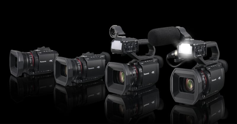 A group of four black Panasonic 4K professional video cameras is displayed against a black background. Two cameras are equipped with microphones and lights, while the other two are without additional accessories.