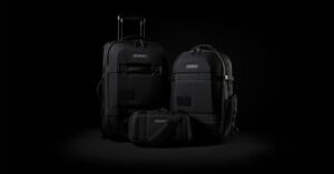 A set of three sleek black travel bags on a dark background: a suitcase with wheels and handle, a large backpack, and a small shoulder bag. They feature durable materials and a minimalist design with subtle branding.