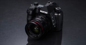 A black Pentax K-3 Mark III camera with a wide-angle lens is placed on a dark, reflective surface. The camera's brand name is prominently displayed, and various dials and buttons are visible on its body.