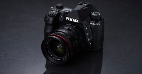 A black Pentax K-3 Mark III camera with a wide-angle lens is placed on a dark, reflective surface. The camera's brand name is prominently displayed, and various dials and buttons are visible on its body.