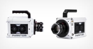 Two white Phantom high-speed cameras are shown against a plain background. Both cameras feature a compact design with black lenses and visible cooling vents. They have sturdy handles on top for easy carrying.