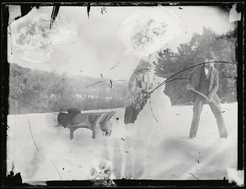 A vintage photograph shows a person in a coat and hat building a snowman, while another person in a suit and hat stands nearby holding a snowball. A wooden wheelbarrow is in the snowy foreground, and trees are in the background.