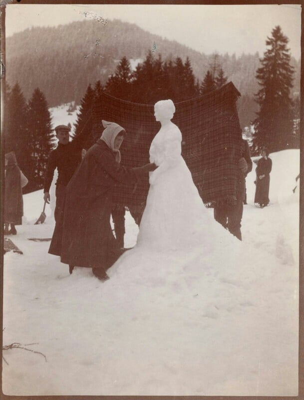 A vintage photograph shows a person sculpting a snowwoman with a shawl on a snowy hillside. A few others stand nearby, and a backdrop of tall trees is visible.