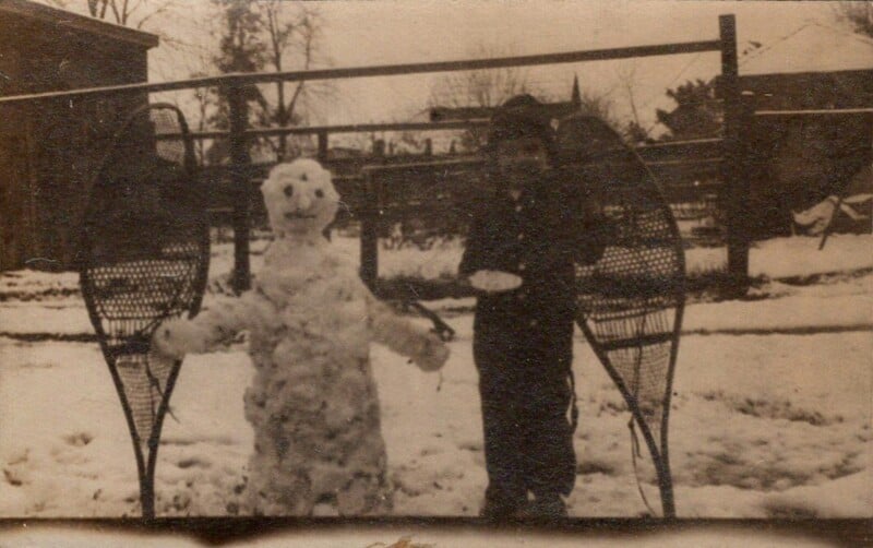 Vintage photo of a child standing next to a snowman with large snowshoes leaning on it. In the background, leafless trees and a fence can be seen. The ground is covered with snow. The image has a sepia tone.