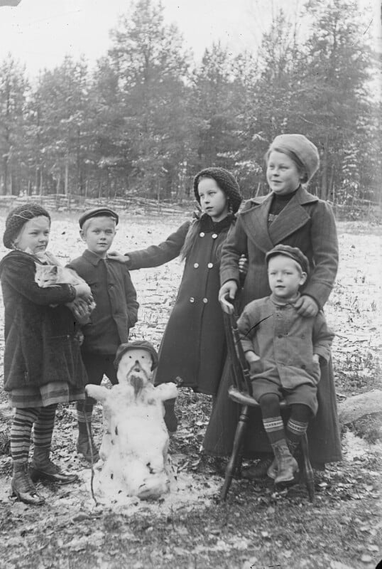 A group of five children wearing winter clothing stands and sits around a small snowman in a field with sparse snow. Four children are standing, and one is seated on a chair. Trees are visible in the background.