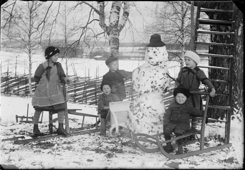 Five children bundled in winter clothing stand around a snowman with a hat, set outdoors near a wooden fence and trees. Two of the children sit on sleds, and there's a snowy landscape in the background.