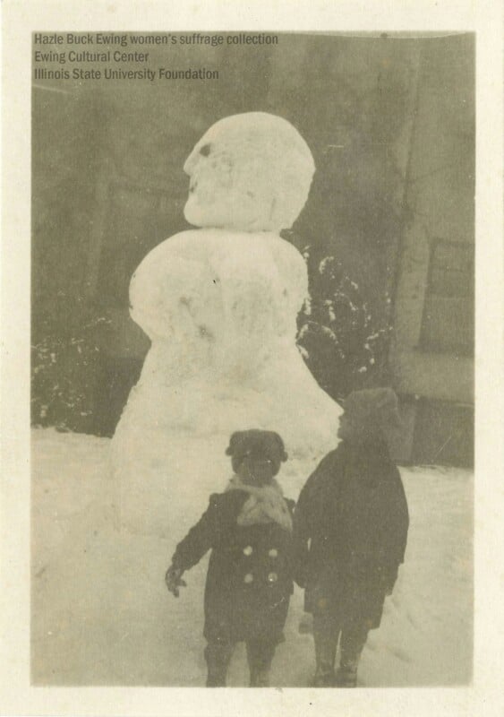 Two children stand in front of a large snowman. The snowman has a rounded body and head, with roughly shaped facial features. The scene is set outdoors, with snow visible on the ground.