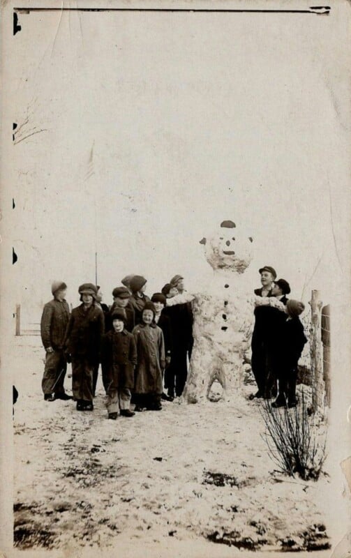 A vintage black-and-white photo shows a group of people, including children and adults, standing next to a large snowman. The snowman has a hat and buttons, and the ground is covered in snow. The group appears to be posing happily.