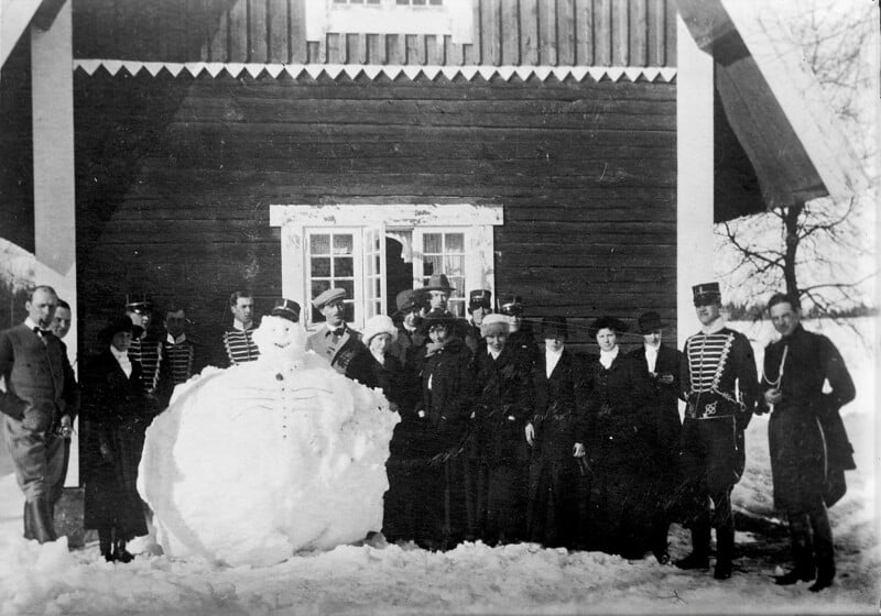 A group of people wearing vintage clothing stands next to a large snowman in front of a wooden house. The group appears to be from an older era, with hats and long coats. Snow covers the ground and roof. The scene looks festive and wintery.