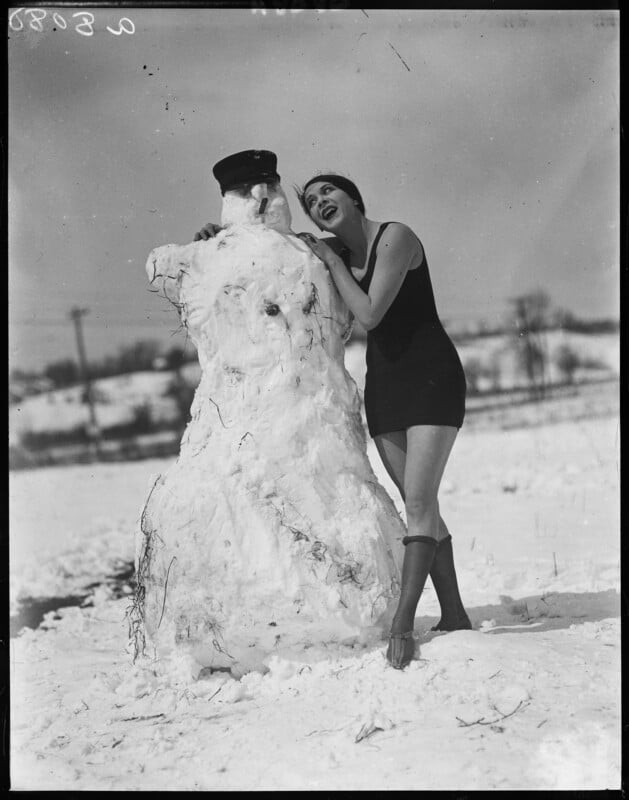 A woman in a sleeveless black outfit and heeled boots stands joyfully next to a snowman wearing a cap. The snowy landscape is visible in the background under a clear sky.