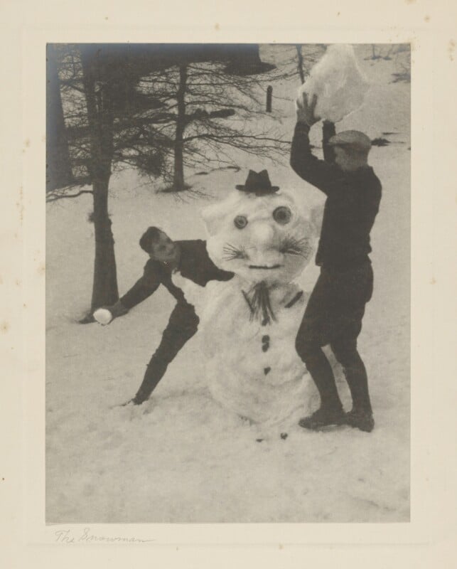 Two people build a snowman in a snowy landscape. The snowman has a hat, eyes, a nose, and a scarf. One person is holding a snowball, and the other is placing snow on the snowman's head. Trees are in the background.