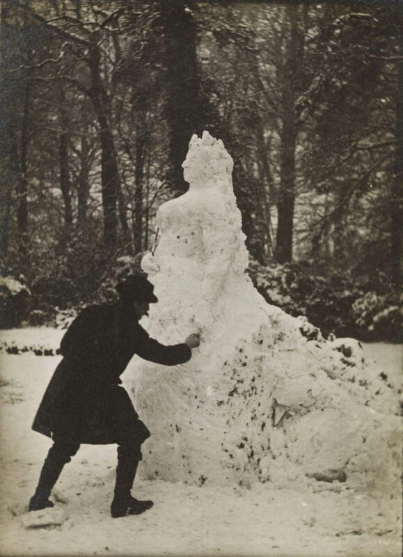 A person sculpting a large, detailed snowwoman in a snowy forest. The person is wearing a dark coat and hat, bending forward while working on the sculpture. Trees covered in snow are visible in the background.