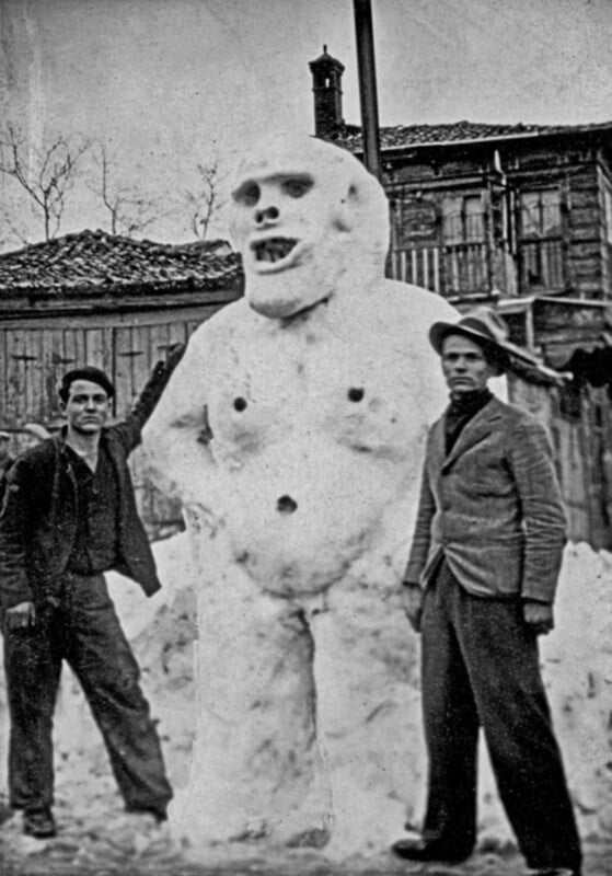 Two men stand beside a large snowman resembling a humanoid figure with pronounced features, including carved eyes and a mouth. The background shows rustic wooden buildings and barren trees. The photo appears vintage, adding to the scene's nostalgic feel.